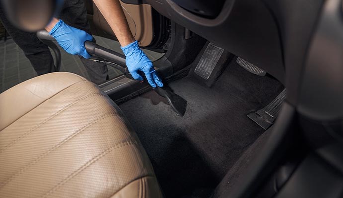 Technician in blue gloves vacuuming the black floor carpet of a vehicle interior near the leather seats