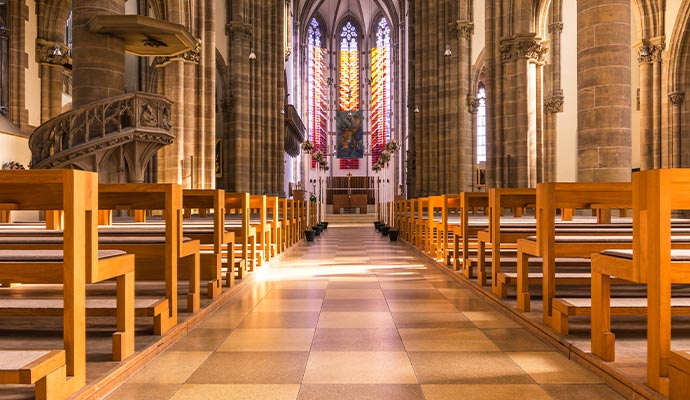 The brightly lit interior aisle of a large church with rows of wooden pews and tall stained glass windows