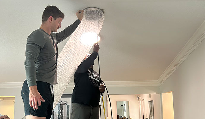 Technicians vacuuming a ceiling air duct with a large vacuum hose.