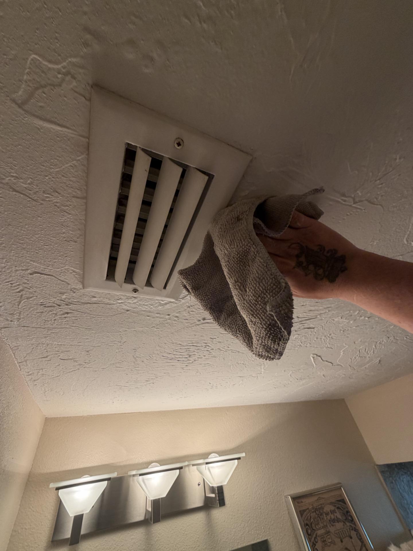 Technician wiping down a ceiling HVAC air vent register to clean condensation staining and mold growth above a bathroom vanity