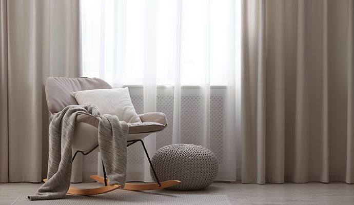 A modern rocking chair positioned next to a bright window covered by clean drapery
