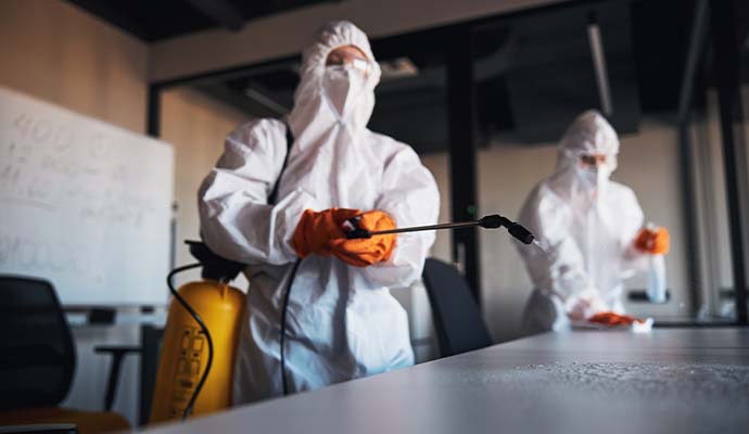 Technicians in protective suits spraying and wiping down office tables with disinfectant