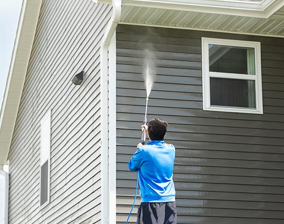 A man in a blue shirt performs soft washing on the side of a house to remove dirt and mildew